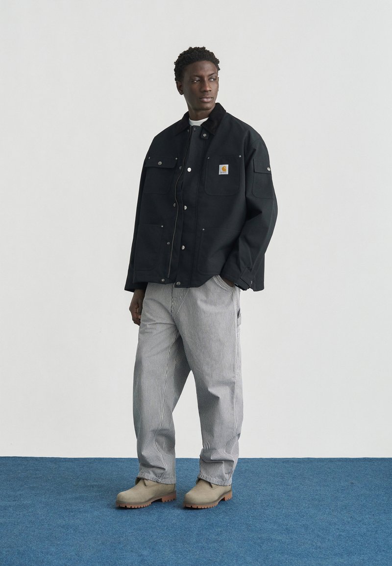 Man wearing black buttoned jacket, striped loose pants, and beige boots standing on blue carpet looking to the side.