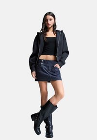 Zwarte zip-up hoodie, aansluitend zwart topje, donkere spijkerrok met knoopaccenten, en kniehoge zwarte leren laarzen met gespdetail.