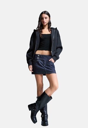 Crna zip-up dukserica, uska crna majica, tamnoplava traper suknja s dugmadi, i crne kožne čizme do koljena s detaljima kopčanja.