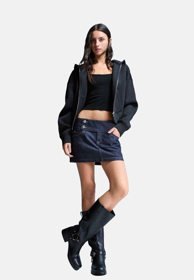 Zwarte zip-up hoodie, aansluitend zwart topje, donkere spijkerrok met knoopaccenten, en kniehoge zwarte leren laarzen met gespdetail.
