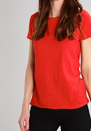 Žena s dlouhými hnědými vlasy, oblečená v jednoduchém červeném tričku s krátkým rukávem a černých kalhotách, stojící na pozadí světle šedé barvy.