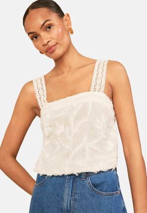 Jonge vrouw met gevlochten haar draagt een mouwloze witte gestructureerde top met kanten bandjes en een blauwe high-waist spijkerbroek, poseert met hand op heup.