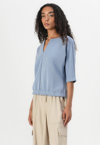 T-shirt azul claro de mangas curtas com decote em V, bainha franzida e tecido suave. Combinado com calças cargo bege com bolsos laterais.