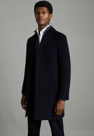 Marineblauer Wollmantel mit strukturierter Silhouette, Reverskragen und drei vorderen Knöpfen. Über einem weißen Hemd getragen, präsentiert er einen formellen Look.