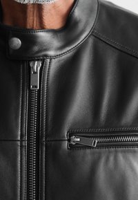 Schwarze Lederjacke mit hohem Kragen, zentralem Reißverschluss und einer kleinen Reißverschlusstasche auf der Brust, die eine glatte Textur und Nähdetails präsentiert.