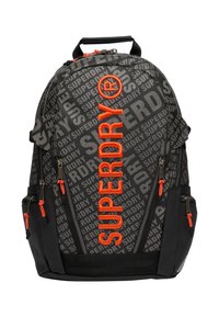 Czarny plecak z napisem "SUPERDRY" w kolorze pomarańczowym, posiadający wiele zamykanych na zamek kieszeni, ergonomiczną rączkę oraz regulowane paski.