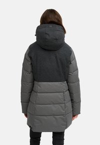 Padded jakna u tamnosivoj boji s teksturiranim vunenim gornjim dijelom i glatkim, pufastim donjim dijelom. Ima veliku kapu i suptilni brending na leđima.