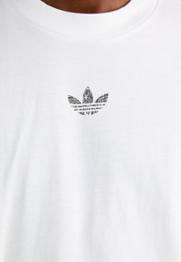 Tricou alb din bumbac, cu un mic logo texturat în formă de frunză neagră pe piept. Guler rotund confortabil, mâneci scurte.