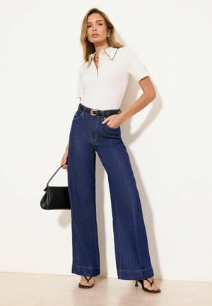 Kvinde iført hvid kortærmet top og højtkalvede viddeblå jeans, der holder en sort håndtaske og står mod en hvid væg.