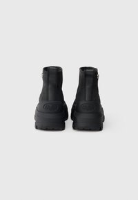 Fekete, vastag talpú platform csizma pár hátulról, hurkos húzófüllel és dombornyomott márkalogóval a sarkán.