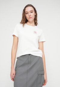 Λευκό βαμβακερό μπλουζάκι με κοντά μανίκια και ένα μικρό, κόκκινο τυπωμένο σχέδιο στο αριστερό στήθος, συνδυασμένο με γκρι cargo φούστα.