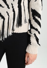 Pletený svetr v krémové a černé barvě, s třásněmi podél spodního okraje a rukávů, s geometrickým vzorem a žebrovanými manžetami.