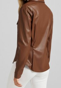 Camicia in eco-pelle marrone con colletto e due tasche sul petto. Presenta maniche lunghe e un orlo leggermente curvo.