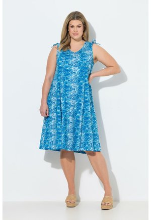 Kobieta ubrana w niebieską, bezrękawną sukienkę z nadrukiem do kolan z wiązanymi ramiączkami, stoi z jedną ręką na biodrze i nosi beżowe sandały na platformie.