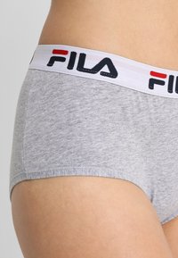 Sivi bombažni šort s belim elastičnim pasom, na katerem je napis "FILA" v črni in rdeči barvi. Gladka tekstura, klasičen kroj, ki pokriva celotno područje.