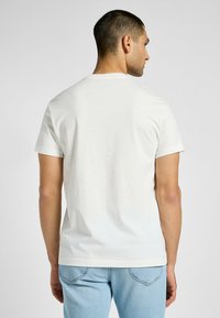 Biała bawełniana koszulka z krótkim rękawem i okrągłym dekoltem, zaprezentowana od tyłu. Gładka tekstura, dopasowany krój. Widoczne jasnoniebieskie dżinsy.