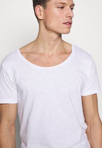 Weißes T-Shirt mit kurzen Ärmeln und weitem Ausschnitt. Hergestellt aus leichtem, strukturiertem Material mit einer lässigen Passform. Glatte Oberfläche, minimalistisches Design.
