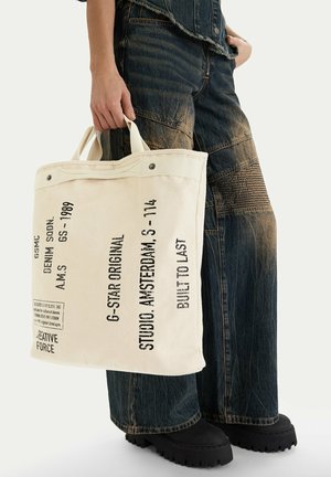 Persona che tiene in mano una borsa tote di tela color crema con testo nero, indossa jeans larghi scuri sbiaditi e scarpe nere chunky