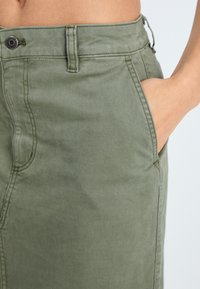 Cotton canvas skirt in olive green, featuring a front pocket, visible stitching, and a button closure at the waist. Smooth texture.