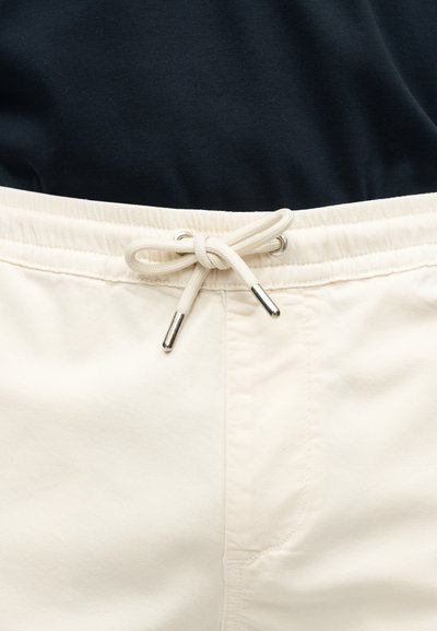 Gros plan sur un pantalon de couleur crème avec une taille à cordon noué et une chemise noire rentrée à l'intérieur.