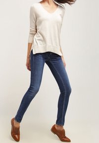Pull beige clair à col en V, manches longues, associé à un jean slim bleu foncé. Des chaussures marron à lacets complètent la tenue.