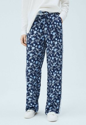 Persoon draagt marineblauwe wijde broek met bloemenprint en zijstrepen, witte sneakers en een los wit langarmshirt.