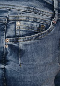 Detailní záběr na kapsu modrých denimových džín s nýty a podrobným žlutým prošíváním, který ukazuje strukturu látky a vybledlá místa.