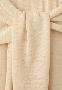 Tissu beige tricoté noué en un nœud lâche, montrant une texture douce et des détails de couture fines.