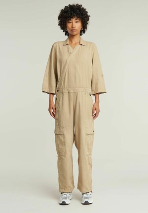 Une combinaison utilitaire beige avec un design enveloppant, des manches larges, des détails de boutons-pression et des poches cargo, associée à des baskets grises et blanches.