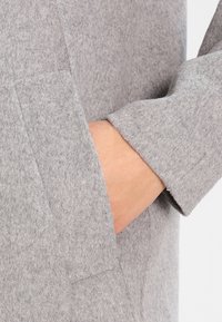 Hånd hvilende inde i lommen på en lys grå uldfrakke med synlig ærmekant og tekstureret stof.