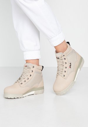 Beżowe buty typu high-top marki Fila z sznurowadłami, noszone z białymi spodniami podwiniętymi na mankiet, przedstawione na białej podłodze i tle.