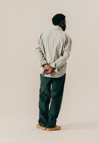 Mann mit Brille und Ohrringen steht mit hinter dem Rücken verschränkten Händen, trägt ein gestreiftes Hemd, dunkle Hose und hellbraune Schuhe vor einem einfarbigen Hintergrund.