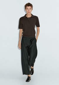 Brązowa dzianinowa koszulka polo z krótkim rękawem, w połączeniu z czarnymi spodniami o wysokim stanie i szerokich nogawkach oraz czarnymi butami. Minimalistyczny design.