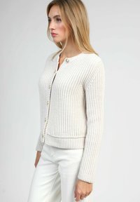 Femme portant un cardigan en maille côtelée blanc avec des boutons, assorti à un pantalon blanc taille haute, présentée de côté sur un fond uni.