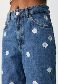 Modré džíny z denimu s bílými vyšívanými sedmikráskami roztroušenými po látce. Mají standardní zapínání na knoflík a design se pěti kapsami.