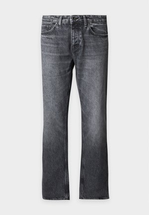 Mand stående med hænderne i lommerne iført beige lynlåshættetrøje, hvid t-shirt, løstsiddende mørke jeans og hvide sneakers på ensartet baggrund.