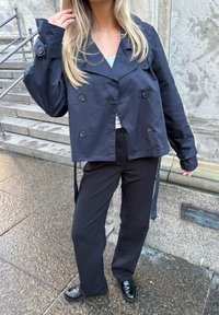 Tamno plavi skraćeni dublje zakopčani kaput s velikim dugmadi, nosi se preko svijetle bluze i crnih hlača, uz sjajne crne cipele.