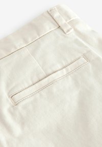 Beige puuvillahousut, joissa on tasainen takatasku ja sileä rakenne, sekä näkyvä vyölenkki ylhäällä varmistamaan turvallisen istuvuuden.