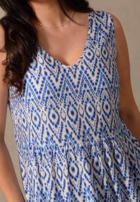Ärmlös klänning med V-ringning, med ett vitt och blått geometriskt mönster, en rynkad midja och en mjuk tygkänsla.