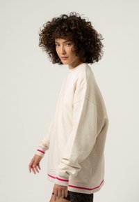 Túlméretezett bézs pulóver rózsaszín csíkokkal a karkivágón és az alján; kerek nyakú és ejtett vállú. Az anyag lágy érzetű.