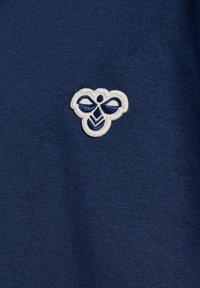 Mörkblå t-shirt i bomull med en vit broderad logotyp i form av ett stiliserat bi med en rosett, mot en bakgrund med jämn textur.