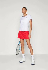 Γυναίκα που στέκεται με λευκή αθλητική μπλούζα, κόκκινη φούστα, λευκές κάλτσες και παπούτσια, κρατώντας μια ρακέτα του τένις απέναντι από έναν απλό φόντο.