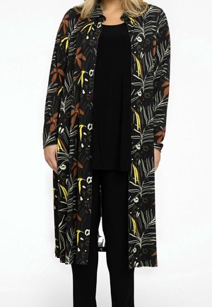 Kvinde iført en lang sort cardigan med orange, gule og hvide blad- og blomsterprint over en sort top og sorte bukser.
