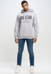 Pelēks sporta krekls ar lieliem melniem burtiem "BIG STAR" un mazākiem burtiem zem tā. Priekšējā kabata, mīksta auduma, apvienots ar ziliem džinsiem un baltām kedām.