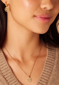 Gouden sieraden met een ronde hanger met een boomontwerp en bijpassende oorringen. Delicate ketting met een textuur van een trui op de achtergrond.