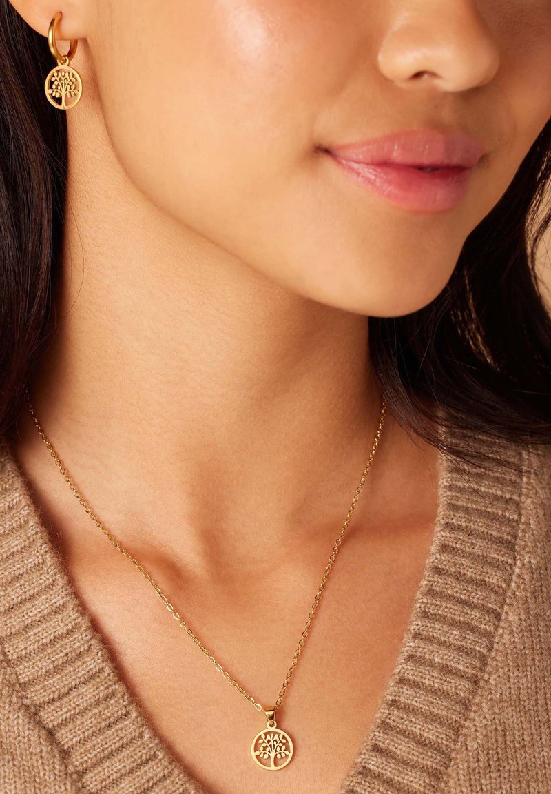 Gouden sieraden met een ronde hanger met een boomontwerp en bijpassende oorringen. Delicate ketting met een textuur van een trui op de achtergrond.