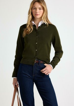 Kvinde iført mørkegrøn knappet cardigan over hvid skjorte med mørkeblå jeans, der holder en brun læderhåndtaske mod en ensfarvet baggrund.