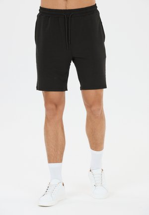 Mand iført sorte shorts med snøre, hvide sokker og hvide sneakers står mod en hvid baggrund.