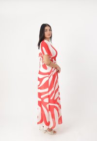 Abito maxi con un motivo astratto rosso e bianco, maniche corte a farfalla, vita aderente e silhouette fluida. Tacchi visibili in basso.