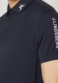 Laivastonsininen pikeepaita puuvillasekoitetta, jossa klassinen kaulus, napillinen edusta ja valkoinen brodeerattu logo kauluksessa ja hihassa.